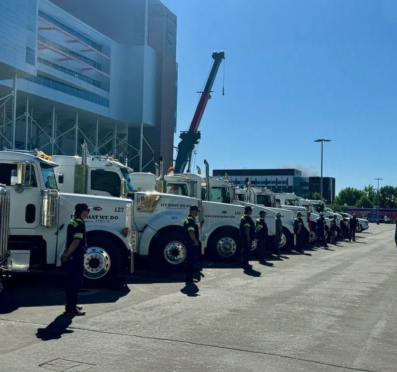 Row of B&W Wrecker Service tow trucks and crane truck lined up with drivers standing beside them