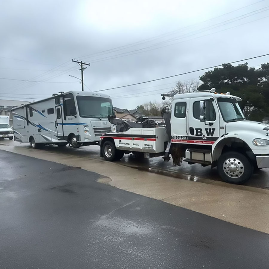 B&W Wrecker Service tow truck towing a Class C RV motorhome on a wet residential street